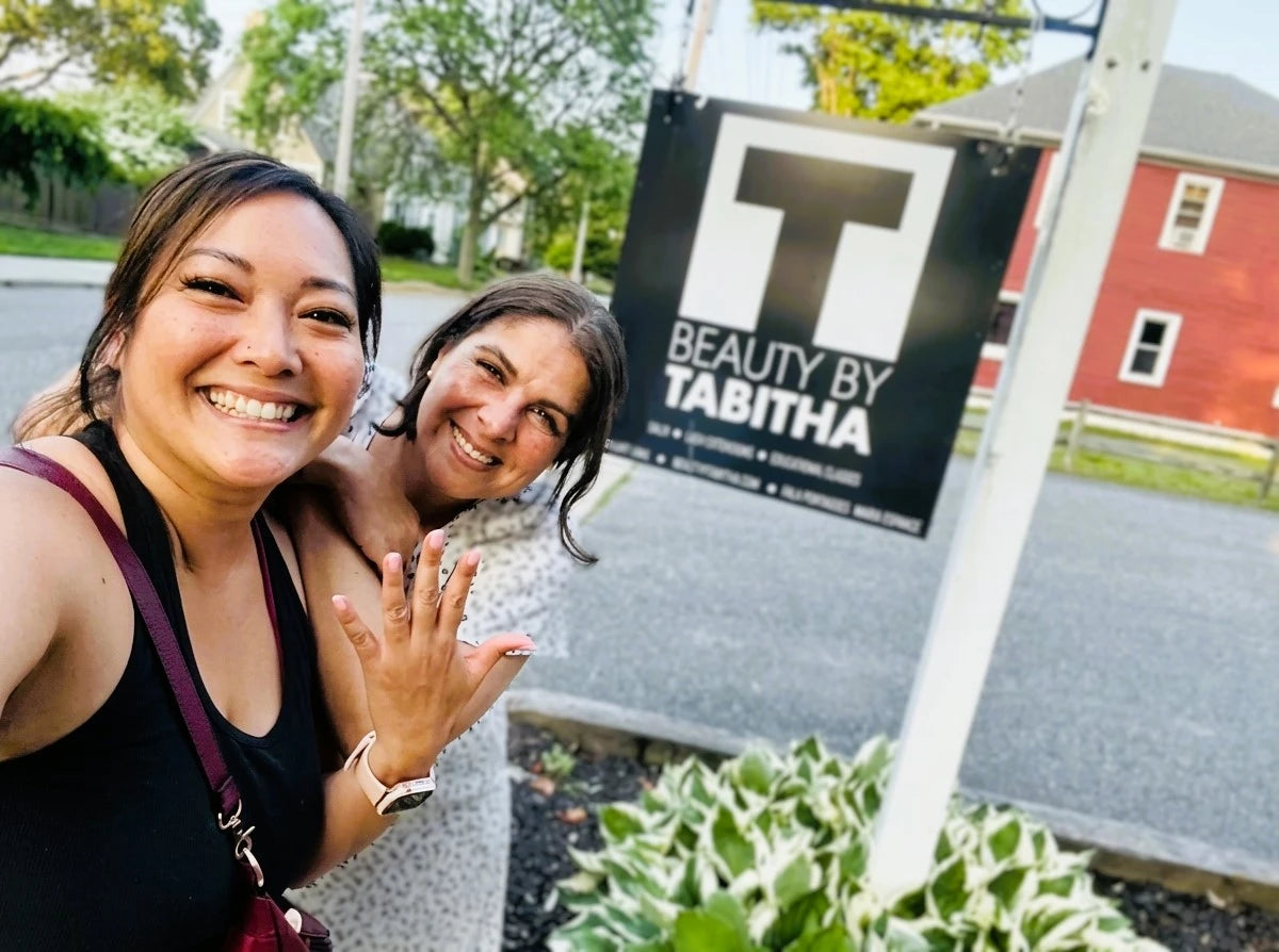 two women in front of a beauty by tabitha sign one woman showing off her nails