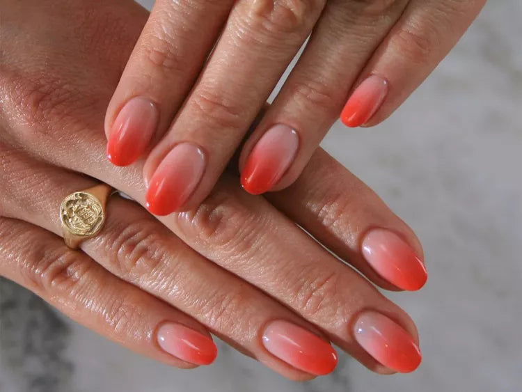 Manicured nails featuring a coral-to-nude ombre design with a gold signet ring.